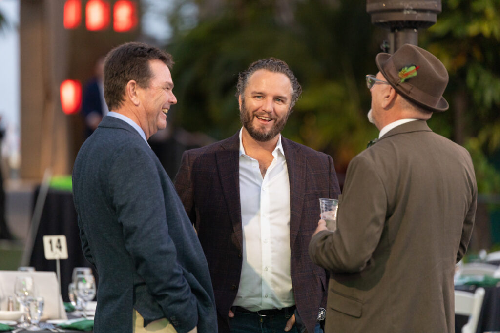 Three men engaged in a lively conversation outdoors at an event, with one facing the camera and the other two with their sides shown. they are well-dressed, standing near a seated dining area.