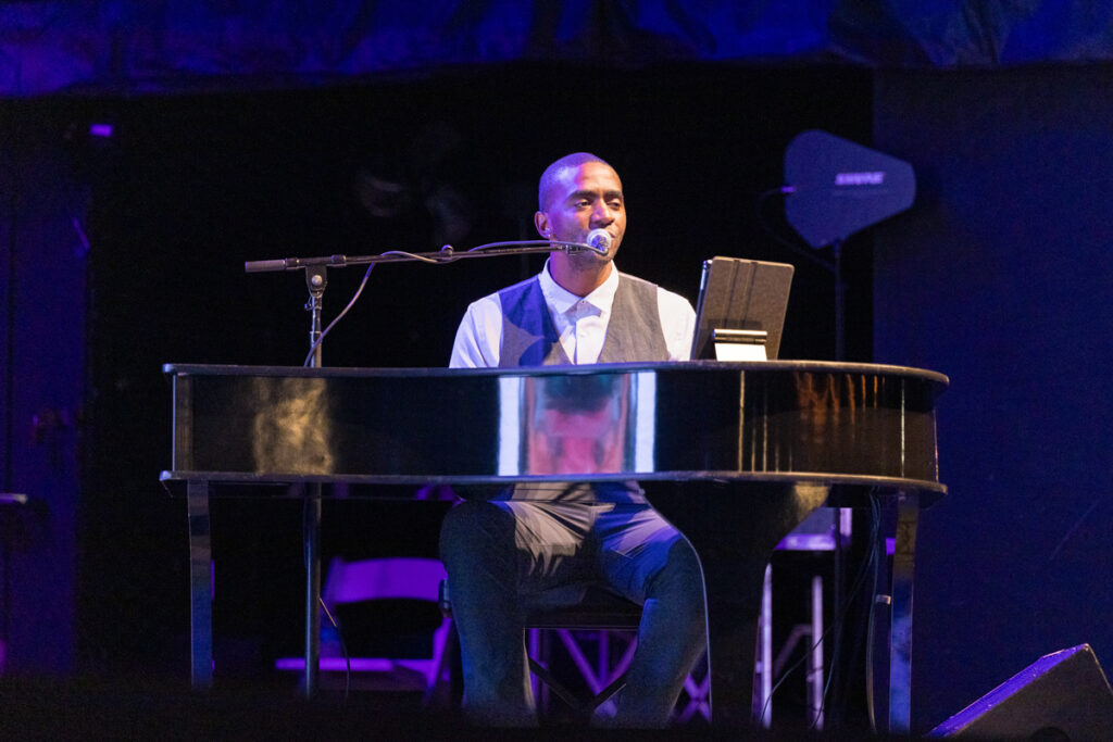 A man sitting at a grand piano under blue stage lighting, performing with a microphone close to his face, with a laptop and music stand nearby.