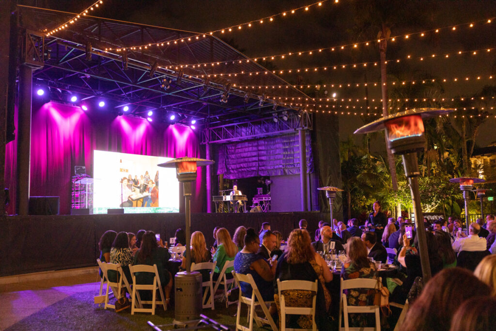 An evening event outdoors with attendees seated at tables, watching a presentation on a large screen under string lights, with stage lighting and heaters visible.