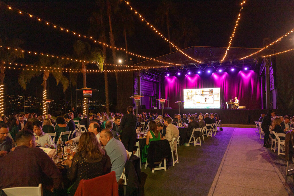 Outdoor evening event with guests seated at tables under string lights, enjoying a live performance on a stage, surrounded by palm trees.