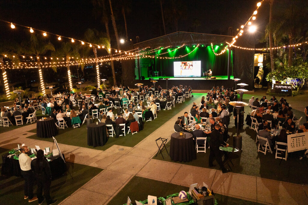 An outdoor evening event with guests seated at tables under string lights, watching a presentation on a large stage and screen. greenery and palm trees enhance the venue, creating a festive atmosphere.