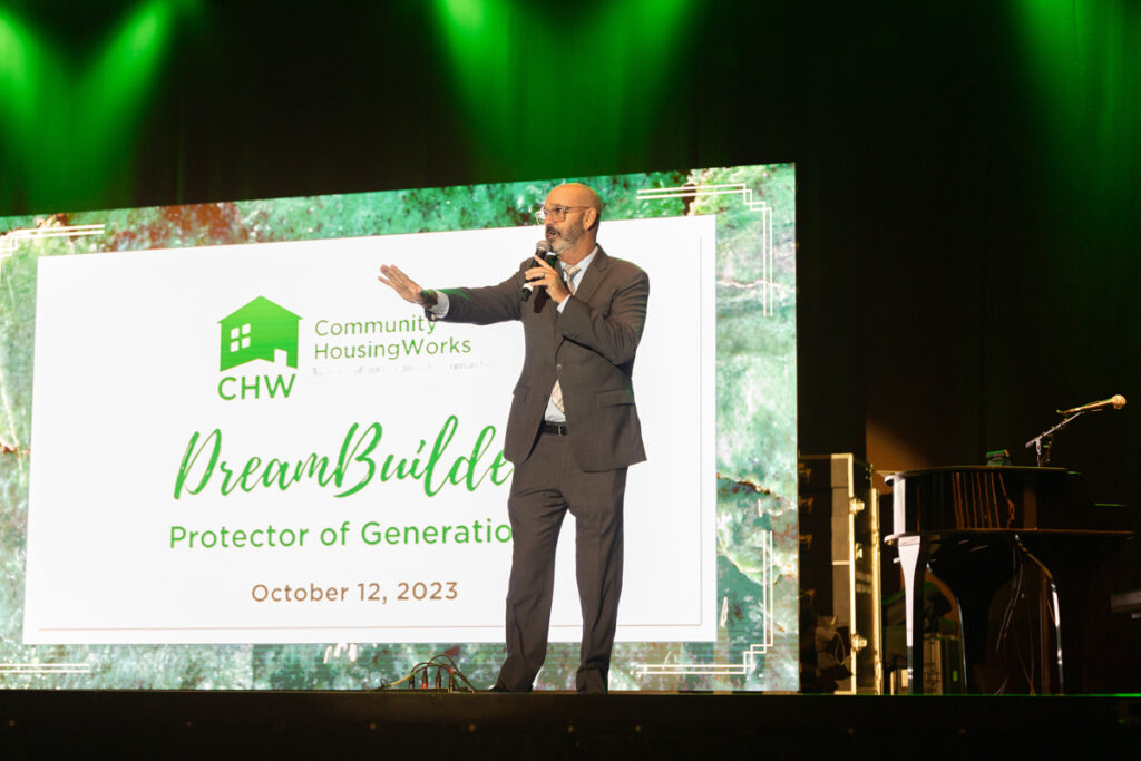 A man gesturing with one hand while speaking at a podium with a large screen behind him displaying the logo and text for "community housingworks dreambuild, protector of generations, october 12, 2023.