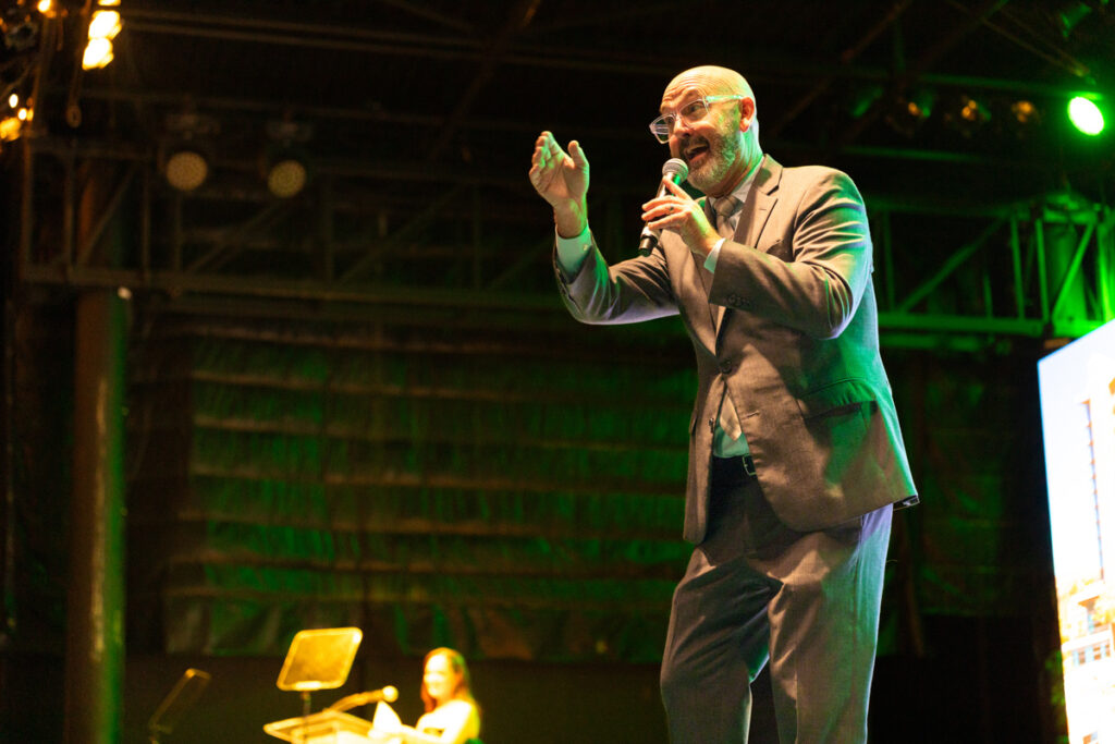 An enthusiastic bald man in a gray suit speaks into a microphone on stage with a brightly lit background, gesturing with his right hand, while a woman in the background looks on.