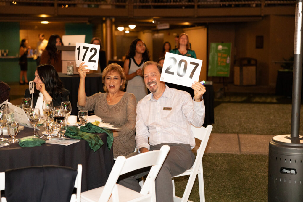 A man and woman seated at a dinner event, joyfully holding up auction paddles numbered 172 and 207, surrounded by other attendees in a dimly lit venue.