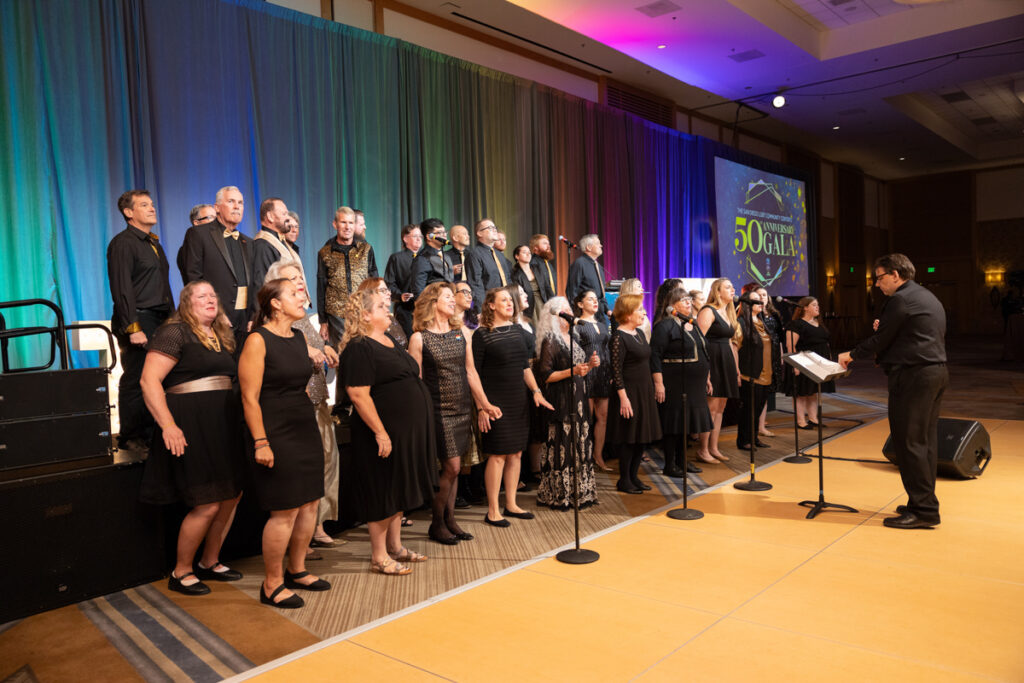 A choir stands on stage in formal black attire with a conductor in front, holding a music folder, during a performance at a 50th anniversary celebration event.