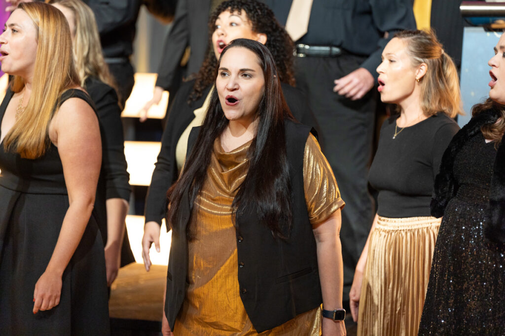 A diverse choir performing on stage, with a woman in a gold blouse and black vest passionately singing in the foreground, surrounded by other choir members in black attire.