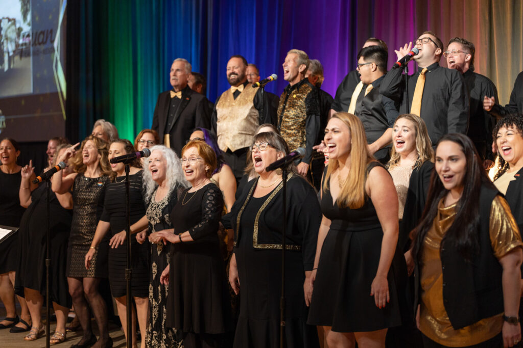 A diverse choir enthusiastically singing on stage during a performance, with vibrant stage lighting in the background. members are dressed in formal black attire with some in gold accents.