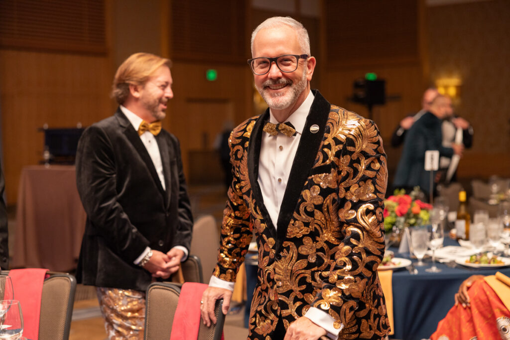 Two men at a formal event, one in a black tuxedo and the other in a unique gold and black patterned jacket, both smiling and standing near a dining table.