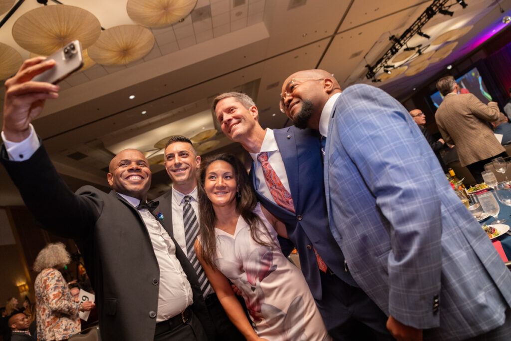 Five people, four men and one woman, take a group selfie at a formal event with a lively background. one man holds the phone. they all smile joyfully at the camera.