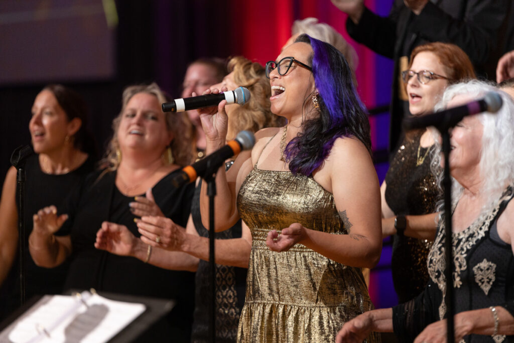 A woman with vibrant purple hair and glasses sings passionately into a microphone, flanked by a choir of women clapping and singing in a concert setting.