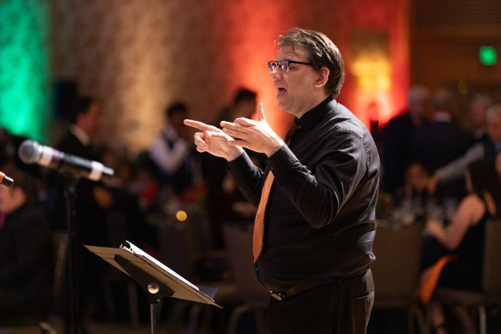 A conductor vigorously directing an orchestra, wearing glasses and a black shirt, in a banquet hall with colorful lighting in the background.