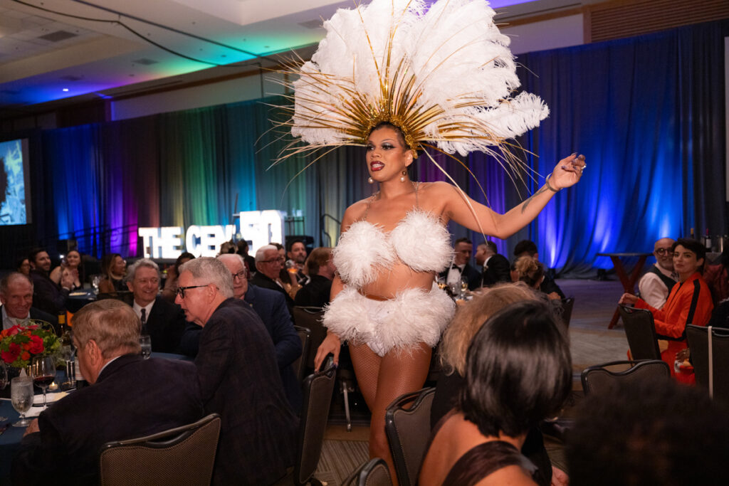 A performer in a striking white feather costume, including a large headpiece, entertains an audience at a gala event, gesturing grandly as attendees watch in appreciation.