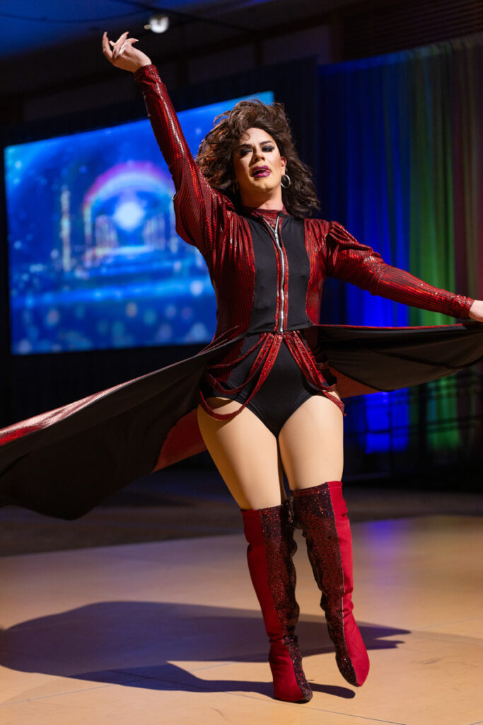 A drag queen performs confidently on stage, wearing a dazzling red and black outfit with thigh-high boots, striking a dynamic pose with arms raised and a beaming smile.