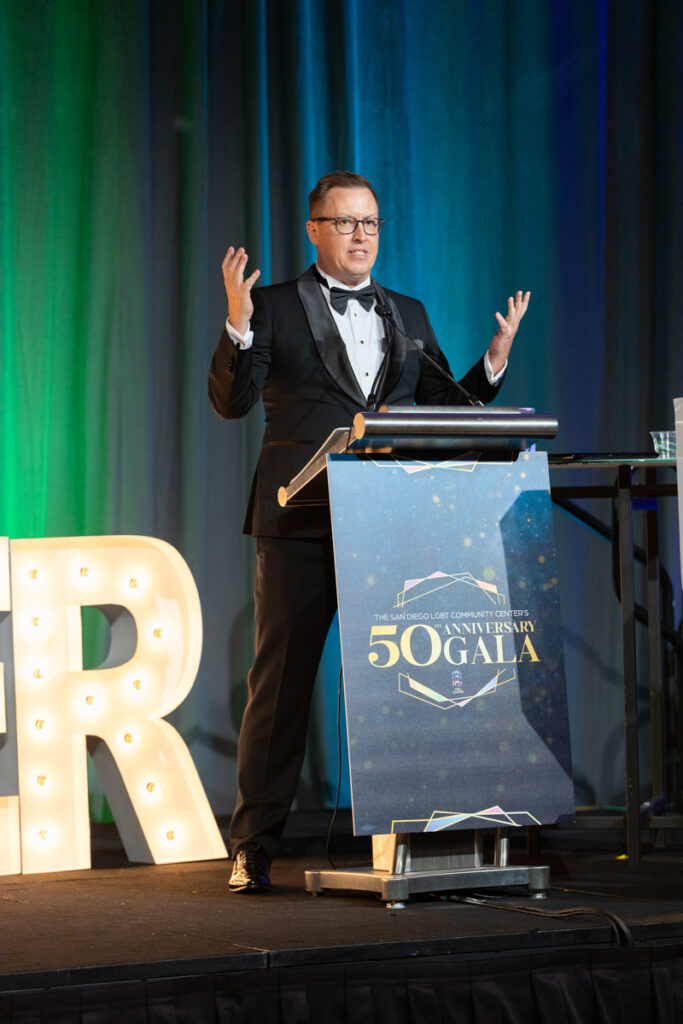 A man in a tuxedo speaks animatedly at a podium with a microphone, during a gala event celebrating a 50th anniversary, with a large lit-up letter "r" on the stage beside him.