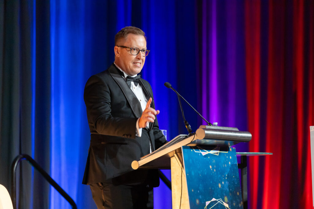 A man in a tuxedo giving a speech at a podium on a stage with a red curtain background. he is holding a remote and there is a microphone in front of him.