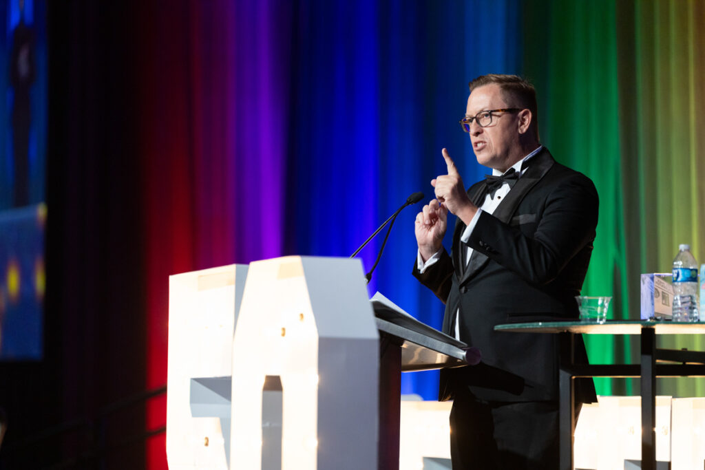 A man in a tuxedo stands at a podium, gesturing with one hand, speaking at an event with a colorful, lit backdrop.