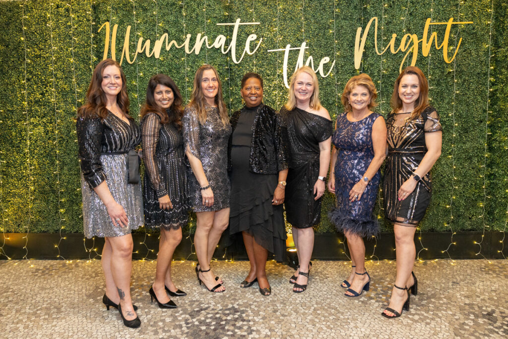 Seven women dressed in elegant, sparkly dresses stand smiling against a backdrop with the phrase "illuminate the night" written in golden cursive, surrounded by a lush green wall.