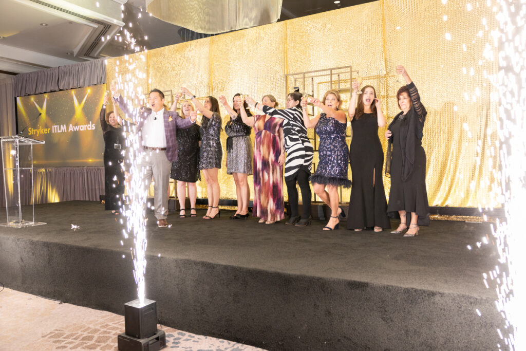 Group of people celebrating on stage with sparklers at a glittering awards event, joyful arms raised, under the "stryker tlm awards" banner.