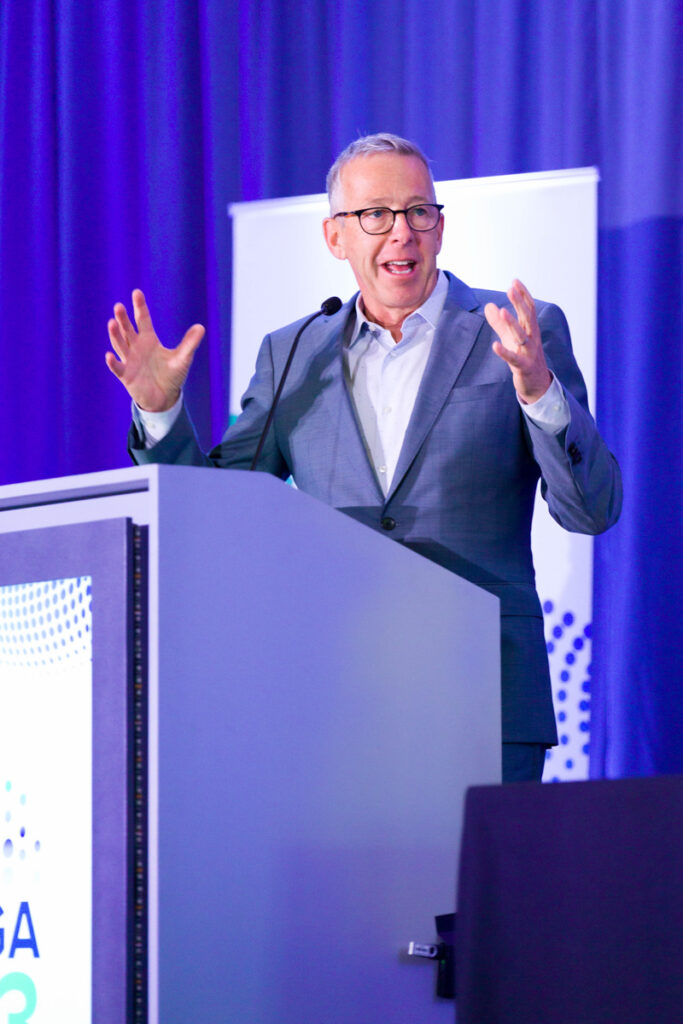 A man in a gray suit and glasses enthusiastically speaking at a podium with a blue banner, in a conference setting with purple lighting. he gestures with his hands mid-speech.