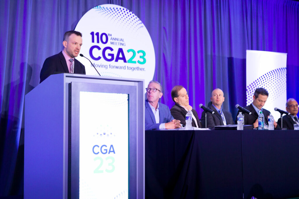 A man speaks at a podium during the 110th annual cga meeting, themed "moving forward together." four other men listen from a panel table against a backdrop with the event's logo.