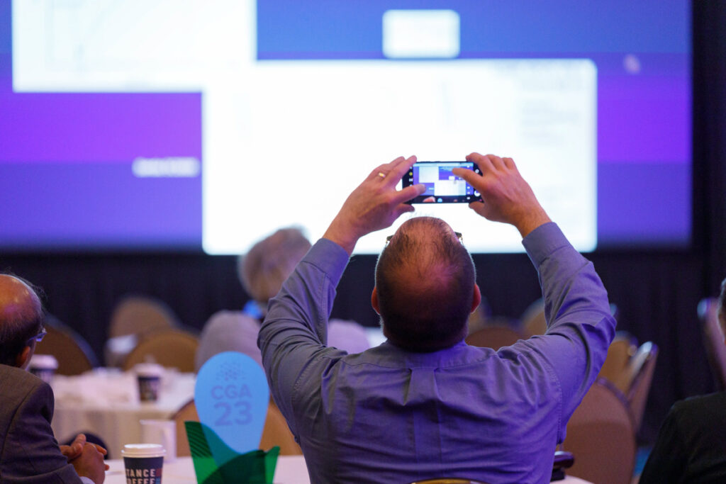 A person at a conference using a smartphone to take a photo of a presentation slide projected on a screen, capturing the moment amidst other seated attendees.