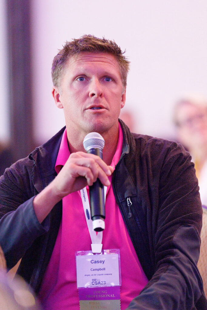 A man with short blond hair, wearing a pink polo shirt and a conference badge, speaks into a microphone during a seminar. he looks attentively to his right, engaging with the audience.