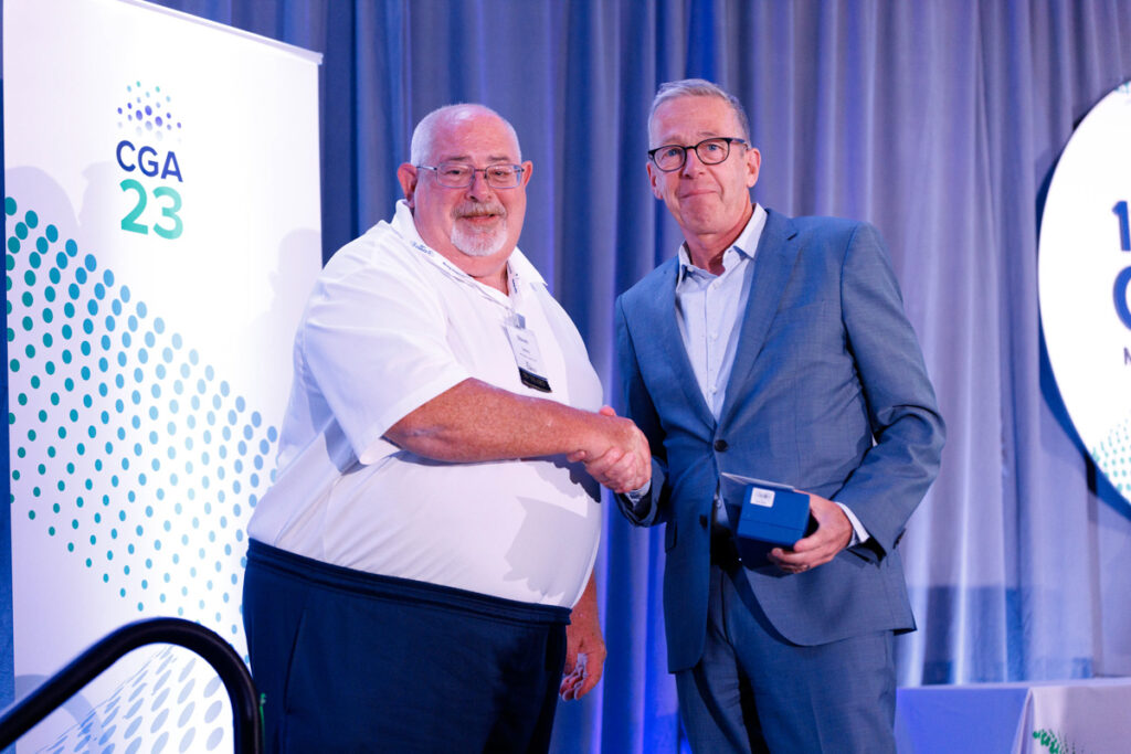 Two men shaking hands on stage at a conference, one receiving a small box from the other, with banners reading "cga 23" in the background.