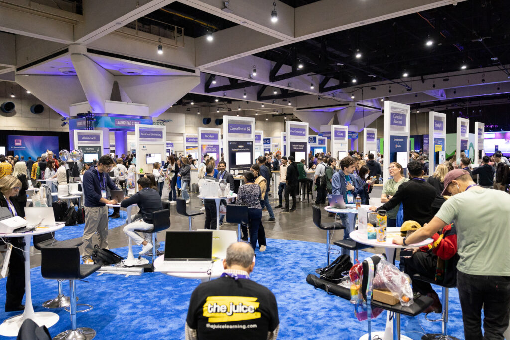 A busy convention hall filled with people interacting at various booths. Attendees are seated and standing, engaging with technology and displays. The atmosphere is lively, with multiple screens and informational signs around the area.