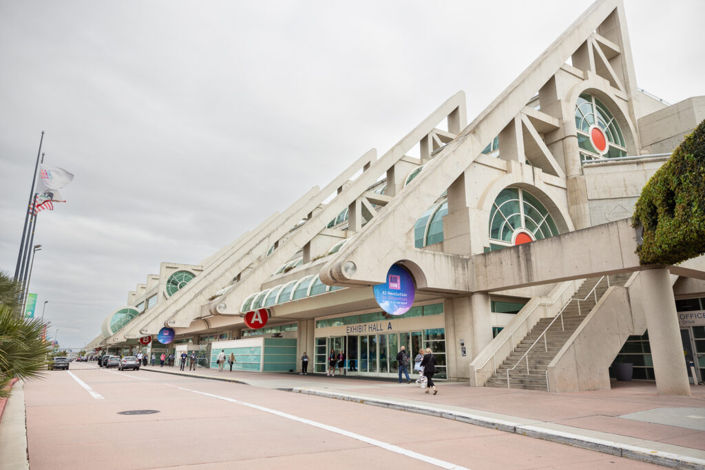 Exterior view of a large convention center with modern architectural design, featuring triangular structures and circular windows. Flags wave on tall poles, and people walk along the sidewalk. A sign for "Exhibit Hall A" is visible.