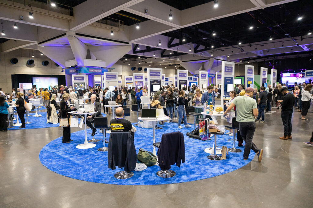 A bustling convention center with attendees gathered around informational booths on a blue circular carpet. People engage in discussions, demonstrating products. Overhead lighting brightens the spacious venue, creating a lively atmosphere.