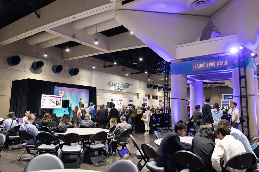 A large indoor event space with many people gathered around a stage area labeled "Launch Pad Stage." There are tables and chairs in the foreground, and attendees are watching a presentation or speaker. The venue is brightly lit.