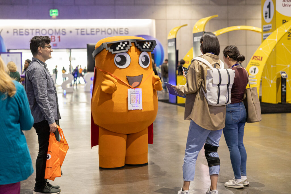 A person in an orange mascot costume with sunglasses interacts with attendees at an indoor event. The setting includes signage for restrooms and telephones, and several people are engaged in conversation.