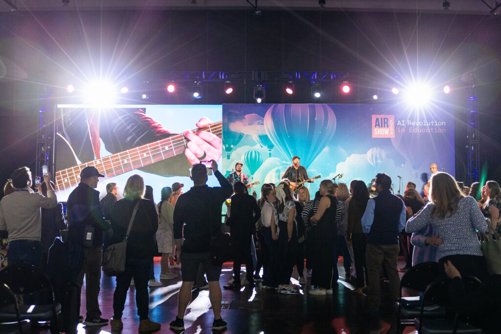 A group of people stand in front of a stage at an event with bright lights. A large screen displays imagery of hot air balloons. The event is titled "AI Revolution in Education.