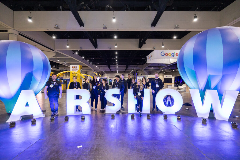 People stand behind large illuminated letters spelling "AR SHOW" at an event. The venue has high ceilings and various booths visible in the background, including one labeled Google. Decorative blue and white balloons are positioned on either side.