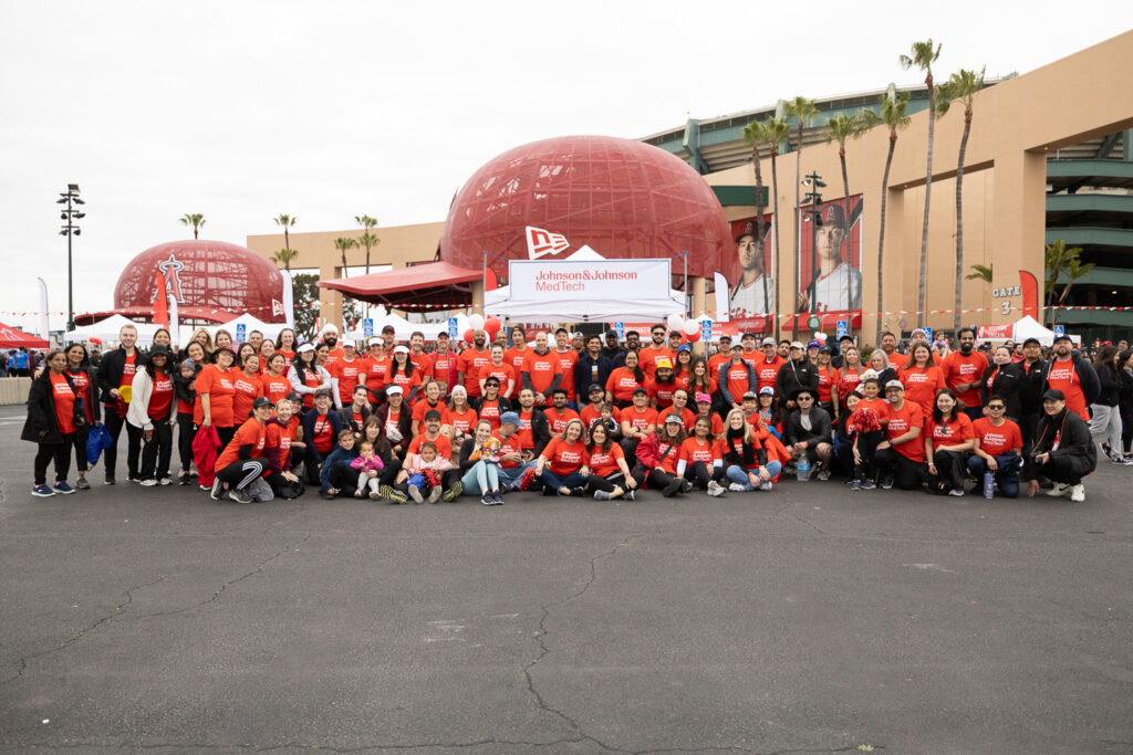 A large group of people, all wearing red shirts, are gathered in front of the Anaheim Angels stadium. They are standing in front of a Johnson & Johnson MedTech banner. The sky is overcast and several palm trees are visible in the background.