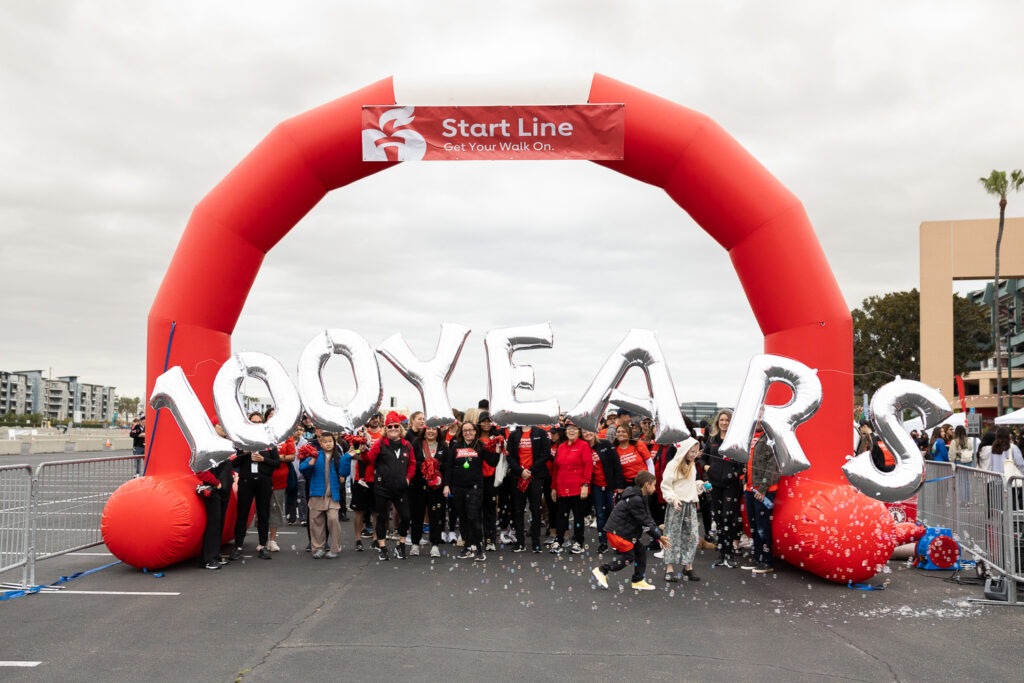 A group of people gathered under a large red arch that reads "Start Line." They hold silver balloons spelling "100 YEARS." Confetti is scattered on the ground, and the event appears celebratory. The sky is overcast.