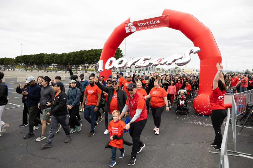 A large group of people begins a charity walk under a red inflatable arch marked "Start Line." Participants wear red shirts, and some hold balloons spelling "100 YEARS." Families with children and diverse ages are visible.