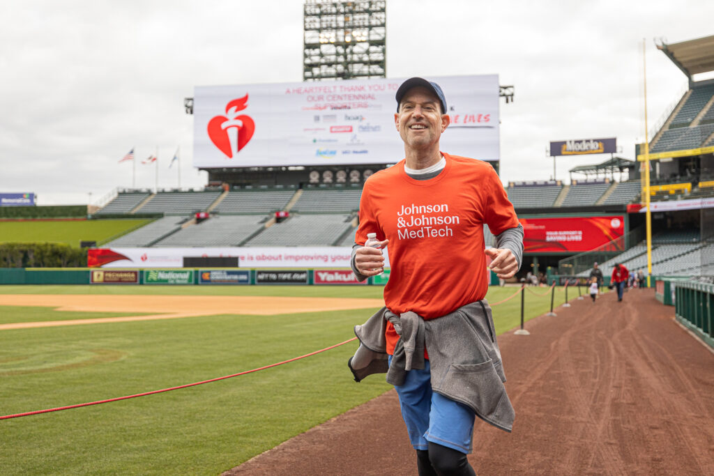 A man in an orange Johnson & Johnson MedTech shirt runs on a stadium track. The stadium has a scoreboard displaying a message and banners with the American Heart Association logo. He's smiling and wearing a cap on a cloudy day.