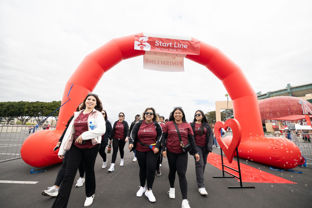 A group of smiling people wearing matching red shirts walks under a large inflatable red arch with "Start Line" printed on it. They appear to be participating in an outdoor event or walkathon. There's a heart decoration on the side.