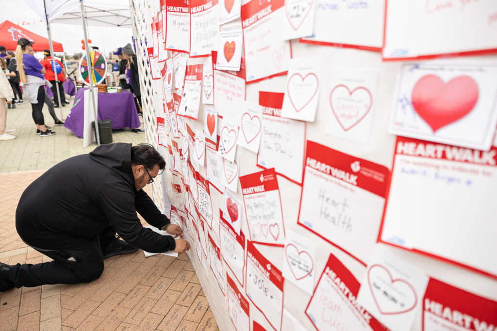 A man kneels to read notes on a wall covered with messages. The notes are heart-themed and part of a health awareness event. Tents and tables are visible in the background, with people milling around.