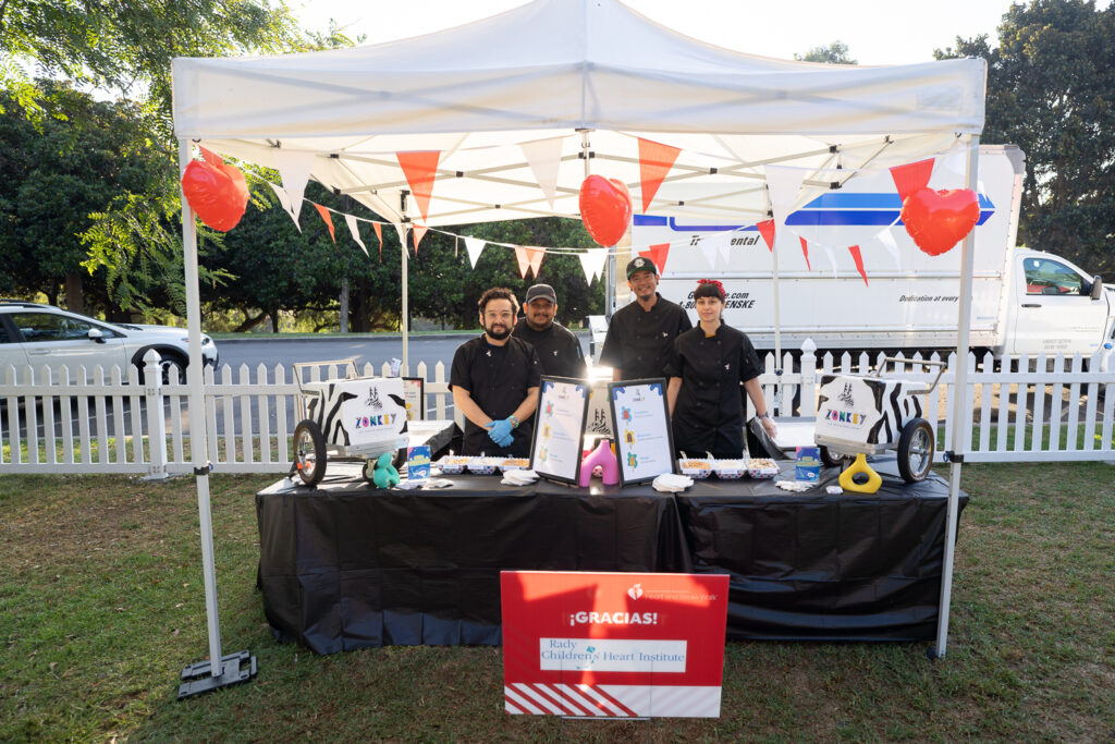 A group of people stands under a white tent with a table displaying food items and menus. The tent is decorated with red balloons and bunting. A sign reads "Gracias!," and a white picket fence and trees are visible in the background.