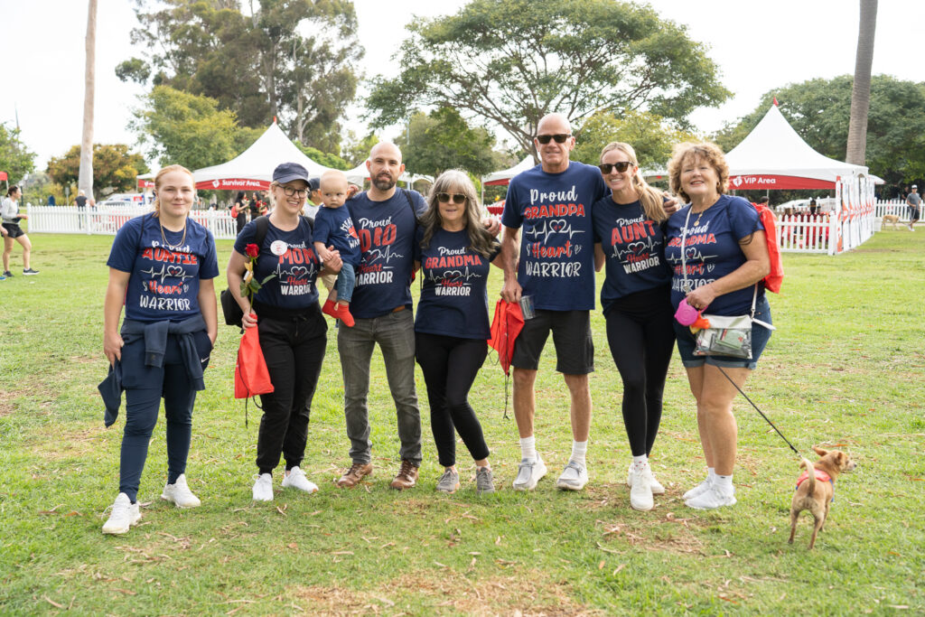 A group of seven people and a small dog stand on grass, smiling and wearing matching navy shirts with red text. Red tents and trees are in the background. Some hold jackets, and one person holds the dog's leash.