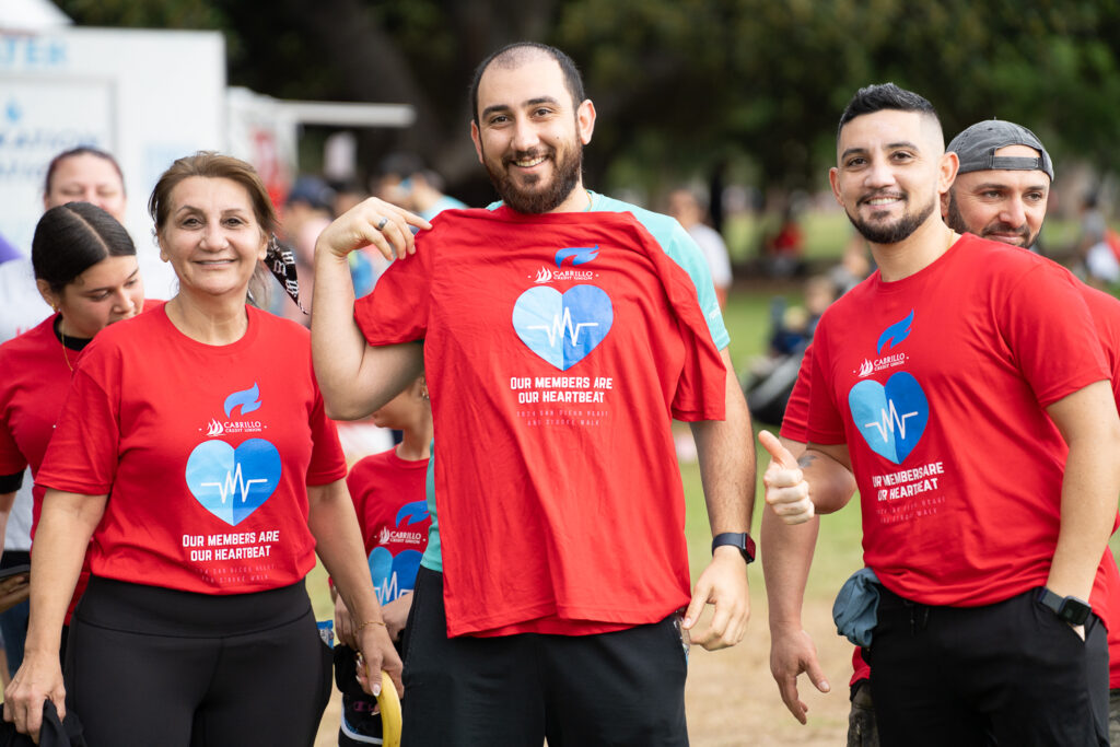 A group of people in red shirts with a heart and text design, smiling at an outdoor event. One man holds an extra shirt. Trees and other participants are in the background.