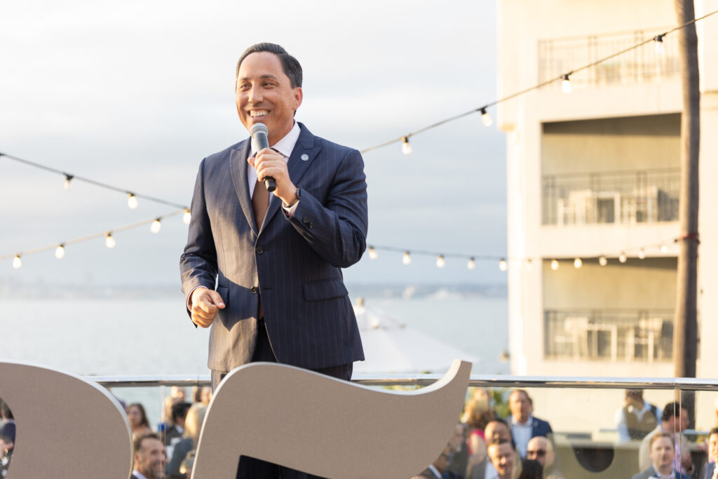 A man in a suit speaks into a microphone at an outdoor event. He stands on a stage in front of a railing with an ocean and buildings visible in the background. String lights are overhead, and a crowd watches in the foreground.