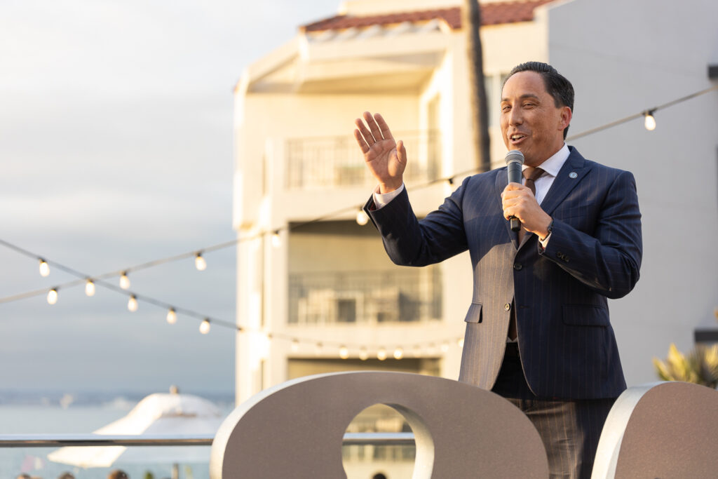 A person in a suit holding a microphone speaks outdoors during a daytime event. String lights and a white building are visible in the background. A large "SD" sculpture is in the foreground.