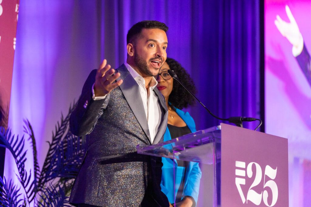 A man in a sparkly suit speaks passionately at a podium with a "25" logo, accompanied by a woman in a blue blazer. The background features purple lighting and plants.