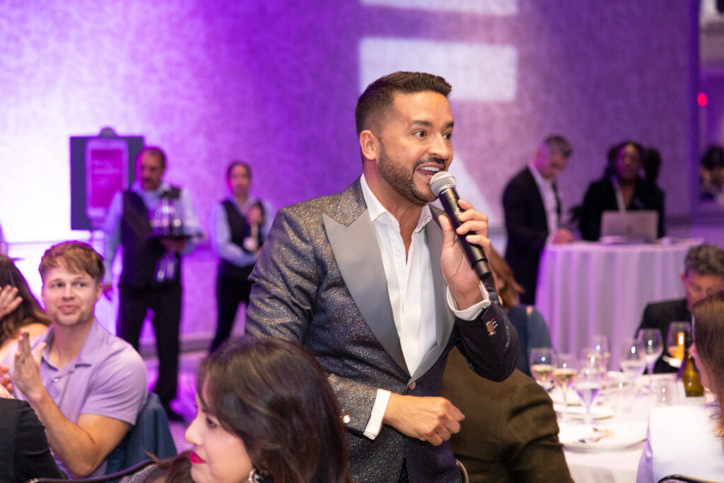 A man in a shiny suit holds a microphone while speaking at a formal event. People are seated at tables with wine glasses and plates. The background has a purple hue with servers and other attendees visible in the room.