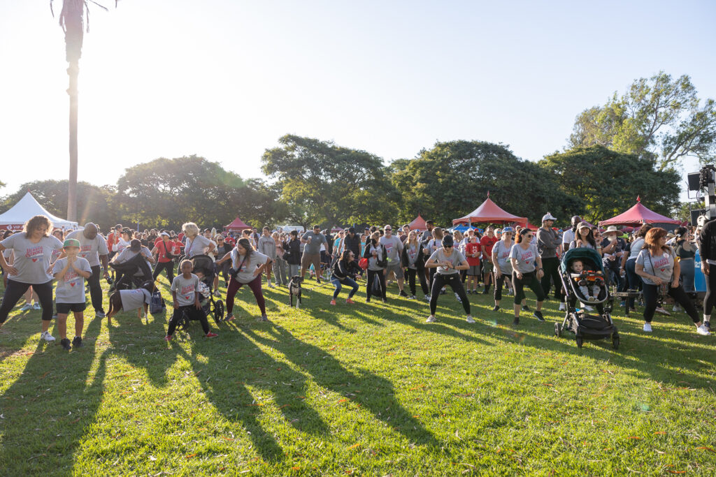 A large group of people, including children, are exercising on a grassy field with trees in the background. Some participants are in workout gear, and there are red canopies around, indicating an outdoor event. Sunlight casts long shadows.