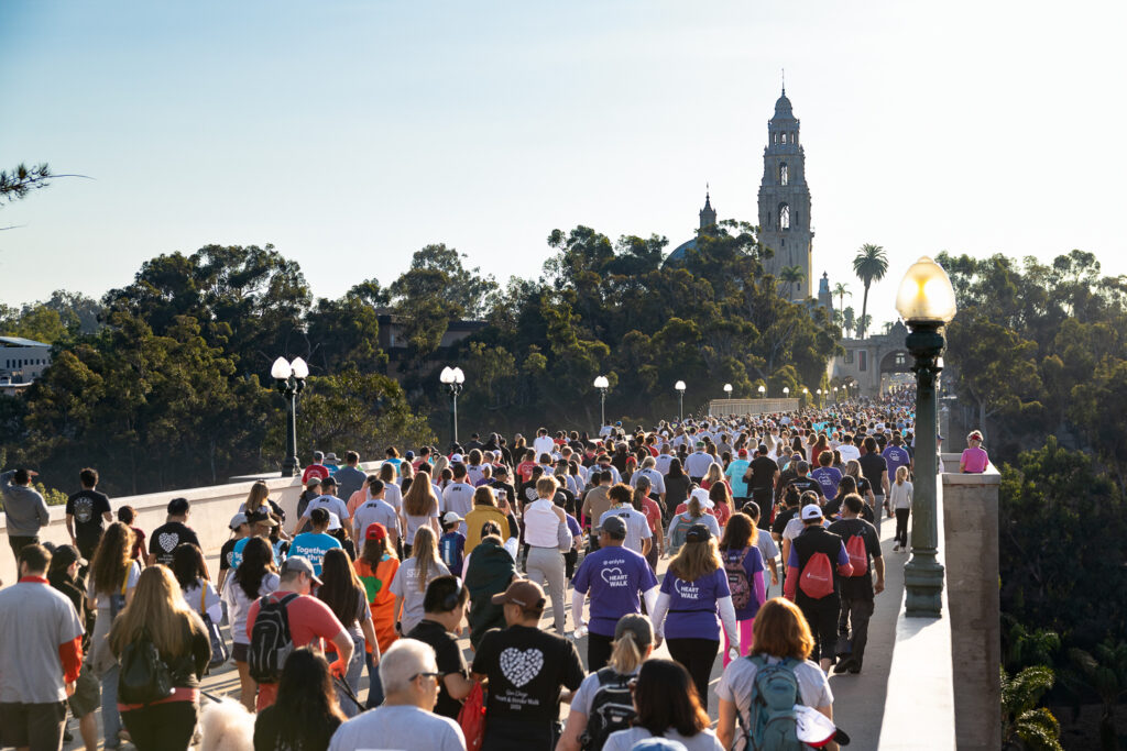 San Diego event Photography Services. A Photo of a large crowd of people, wearing various colorful shirts, walk across a bridge surrounded by trees. In the background, there's a tall, ornate building with a tower. The sky is clear and sunny.