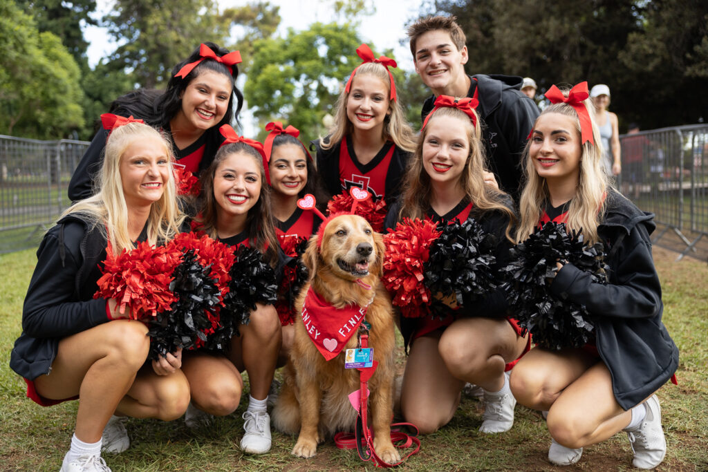 A group of cheerful cheerleaders dressed in black and red uniforms, smiling and crouching around a golden retriever wearing a red bandana. They are outdoors on grass with trees and a fence in the background.